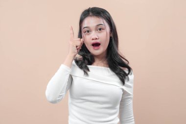 Omzunda beyaz bir bluz olan Asyalı bir kadın parmağını yukarı kaldırıyor. Şaşırmış bir ifadeyle. İlhamın, yeni fikirlerin ve ani farkındalığın sembolü..