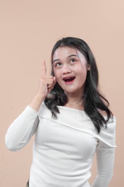 Asyalı bir kadın omuzlarından beyaz bir bluz giyer. Şaşırmış bir yüzle gülümser ve yukarı doğru işaret eder. Fikri, yaratıcılığı, iş konseptini, odaklanmayı ve ilham kaynağını sembolize eder..