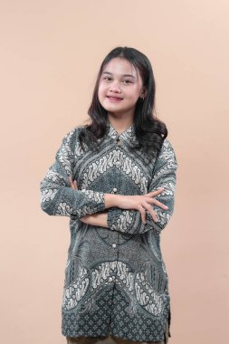 Geleneksel batik elbise giyen kendine güvenen genç bir Asyalı kadın kollarını kavuşturmuş, doğal olarak gülümsüyor, zarafet, güzellik ve profesyonellik gösteriyor..