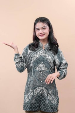 Geleneksel batik tişört giyen Asyalı bir kadın, neşeli bir gülümseme ve açık bir el hareketi ile poz veriyor, modern iş tarzında özgüveni, zarafeti ve olumlu iletişimi simgeliyor..