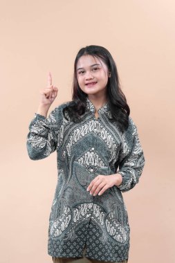 Batik elbiseli Asyalı bir kadın tek parmağını kaldırırken gülümser, kendine güven ve olumlu ifade gösterir, fikri, odaklanmayı ve profesyonelliği sembolize eder..