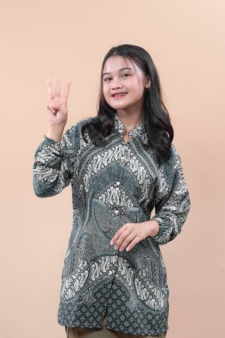 Batik elbiseli Asyalı bir kadın, yaratıcı iş kavramları için güven, cazibe, pozitiflik ve profesyonel stil ifade ederken üç parmağını göstererek gülümsüyor..