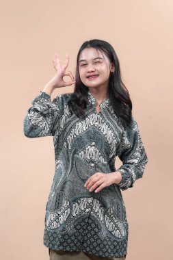 Zarif batik tişört giyen, kendine güvenen genç Asyalı kadın. Tamam el hareketi yapıyor, olumlu bir ifadeyle gülümsüyor. Reklam kavramları için cazibe, profesyonellik ve kültürel gurur gösteriyor..