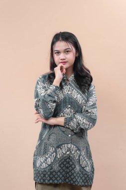 Geleneksel batik tişörtlü Asyalı kadın düşünceli bir ifadeyle poz veriyor, çenesine dokunuyor, özgüvenini ve zarafetini arka planda gösteriyor..
