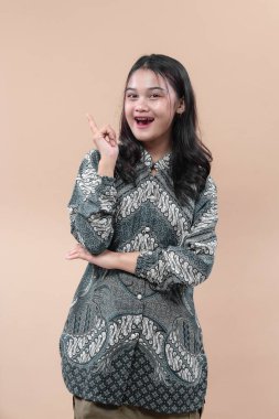 Batik tişört giyen Asyalı bir kadın neşeli bir ifadeyle gülümsüyor, kendine güvenen bir jestle işaret ediyor, bej arka planda izole edilmiş..