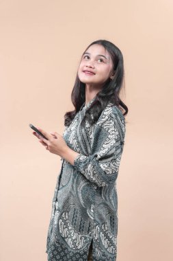 Batik kıyafetli Asyalı bir kadın elinde akıllı telefon tutarken gülümsüyor ve yukarı bakıyor, özgüveni, iyimserliği, kültürel gururu ve modern yaşam tarzını simgeliyor..
