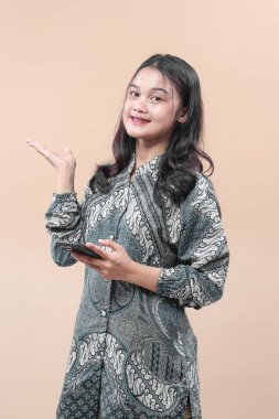 Zarif batik elbiseler giyen Asyalı kadın akıllı telefon tutarken gülümsüyor ve açık el hareketi yapıyor, güven, iletişim, iş, kültür ve modern yaşam tarzını temsil ediyor..