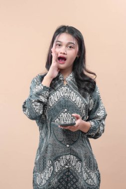 Geleneksel batik giysiler giyen Asyalı bir kadın akıllı bir telefon tutarken haykırarak sürpriz bir jest yapıyor. Duygularını ifade ediyor ve vücut diline güveniyor..