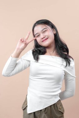 Gözleri kapalı gülümseyen, beyaz bluz ve günlük pantolon giyen, tarafsız bir arkaplanda mutlu bir şekilde poz veren neşeli, genç bir Asyalı kadın..