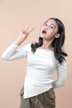 Uzun kollu beyaz bir bluz giyen etkileyici bir Asyalı kadın, tarafsız bir arkaplana karşı güven, neşe ve doğal cazibe gösteren bir barış hareketiyle neşeli poz veriyor..