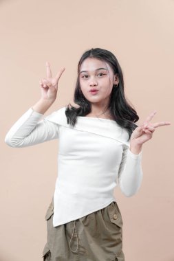 Genç Asyalı kadın neşeli bir yüz ifadesiyle neşeli poz veriyor, beyaz bir bluz ve günlük bir etek giyerken barış eli hareketleri yapıyor, kendine güven, neşe ve gençlik cazibesini gösteriyor,.