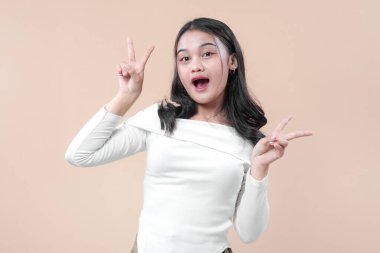 Güzel Asyalı bir kadın neşeli bir gülümseme ve neşeli bir barış eli hareketiyle poz veriyor, beyaz bir bluz giyiyor, kendine güvenini, çekiciliğini, pozitifliğini ve modern yaşam tarzını gösteriyor..