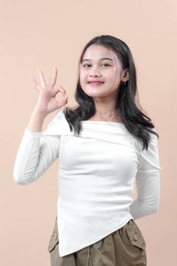 Beyaz, uzun kollu gömlekli Asyalı bir kadın. Nazikçe gülümserken sağ eliyle tarafsız bir arkaplana doğru el hareketi yapıyor..