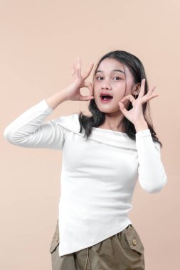 Beyaz, uzun kollu bir gömlek giyen Asyalı bir kadın, iki oyuncu el hareketini yüzüne yansıtarak, tarafsız bir arkaplanda neşe ve güveni ifade ediyor..