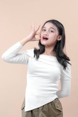 Beyaz uzun kollu tişörtlü Asyalı bir kadın şakacı bir şekilde gülümserken, tarafsız bir arkaplana karşı ağzının yanında el hareketi yapıyor..