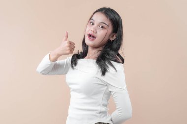 Genç Asyalı bir kadın açık bir yüz ifadesiyle gülümserken başparmağıyla bir jest yapıyor, günlük beyaz kıyafetler giyiyor, pozitiflik, motivasyon ve kendine güven gösteriyor..