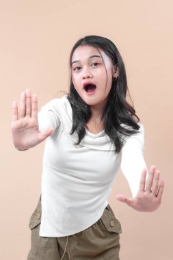 Şaşırmış yüz ifadesine sahip genç Asyalı bir kadın dur işareti olarak ellerini kaldırıyor, günlük beyaz kıyafetler giyiyor, uyarı, korku, reddetme ve ihtiyatı simgeliyor..