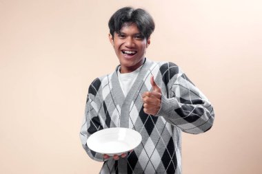 Gri baklava desenli süveter giyen gülümseyen Asyalı bir adam beyaz bir tabak tutuyor ve mutluluk, pozitiflik ve iş hayatı ve kavramları için kendine güveni ifade eden bir baş parmak gösteriyor..