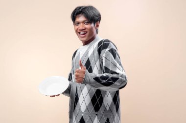 Gri baklava desenli süveter giyen mutlu bir Asyalı, elinde beyaz bir tabak tutarken gülümsüyor ve olumlu, kendine güvenen ve modern yaşam tarzını sembolize eden baş parmağını kaldırıyor..