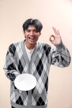 Beyaz bir plaka giyen gülümseyen Asyalı bir adam beyaz bir el hareketi yapıyor, pozitifliği, onayı ve kendine güvenen yaşam tarzını simgeliyor..