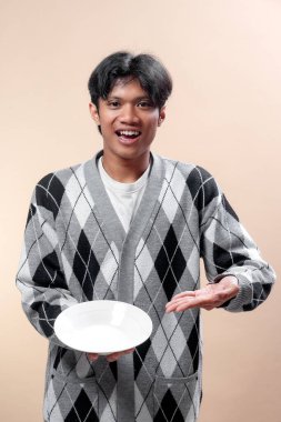 Asyalı bir adam gri bir baklava desenli süveter giyiyor. Elinde boş beyaz bir tabak tutarken gülümsüyor ve eliyle işaret ediyor. Neşeli ve davetkar duyguları ifade ediyor..
