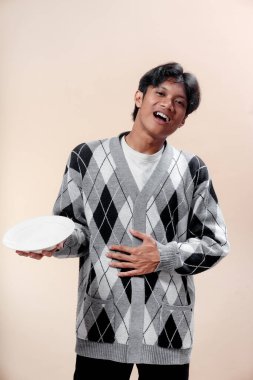 Gri baklava desenli süveter giyen Asyalı bir adam karnına dokunurken boş bir tabak tutuyor ve gülümsüyor, neşeli ve umut dolu bir hareketle açlığını ifade ediyor..