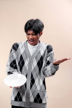 Gri baklava desenli süveter giyen Asyalı bir adam, boş bir tabak tutarken ve bir elini kaldırırken hayal kırıklığına uğramış görünüyor, anlamlı vücut diliyle kafa karışıklığı ve açlık gösteriyor..