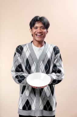 Gri baklava desenli süveter giyen Asyalı bir adam, elinde boş beyaz bir tabak tutarken ışıl ışıl gülümser. Reklam ve yaşam tarzı kavramlarına uygun neşeli ve dostane bir jest yapar..