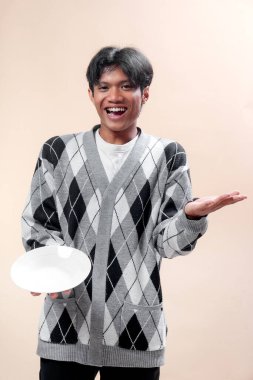 Gri baklava desenli süveter giyen Asyalı bir adam boş bir tabağı tutarken ve elini kaldırırken neşeli bir şekilde gülümsüyor ve dost canlısı vücut diliyle olumlu bir ifade sergiliyor..