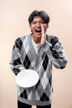 Gri baklava desenli süveter giyen Asyalı bir adam boş bir tabak tutarken gülümsüyor ve elini ağzına dayayıp neşeli ve enerjik bir iletişim hareketi yapıyor..
