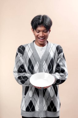 Asyalı bir adam şık gri bir baklava desenli süveter giyerek gülümserken iki eliyle beyaz bir tabak tutuyor ve neşeli ve pozitif bir ruh hali içinde..