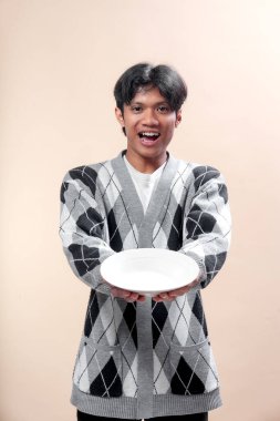Gri bir baklava desenli süveter giyen genç bir Asyalı beyaz bir tabağı ileri doğru tutarken açıkça gülümsüyor, anlamlı ve neşeli bir jest yapıyor..