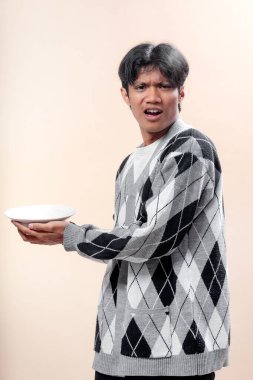 Gri baklava desenli süveter giyen genç bir Asyalı iki eliyle beyaz bir tabak tutuyor. Şaşırmış bir yüz ifadesi ve duygusal tepki gösteriyor..