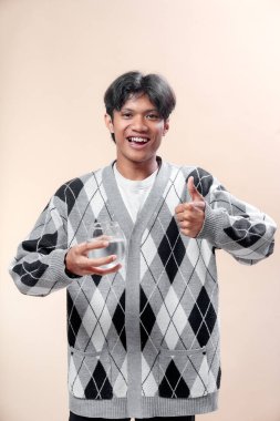Asyalı bir adam gri baklava desenli bir süveter giyer bir bardak su tutarken gülümser ve başparmağını kaldırır, pozitifliğini ve sağlıklı yaşam tarzını ifade eder..