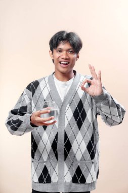 Baklava desenli süveter giyen Asyalı bir adam elinde bir bardak su tutarken gülümsüyor ve el hareketi yapıyor. Kendine güveni, sağlıklı yaşam tarzını ve pozitifliği temsil ediyor..