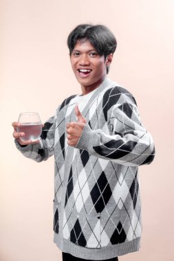 Elinde bir bardak su tutarken gülümseyen gri baklava desenli süveter giyen Asyalı bir adam, onay, sağlık ve pozitif enerjiyi sembolize eden bir jest yapıyor..