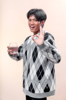 Baklava desenli süveter giyen Asyalı bir adam elinde bir bardak su tutarken gülümsüyor ve el işareti yapıyor. Onayı, sağlığı, pozitifliği ve kendine güvenen yaşam tarzını simgeliyor..