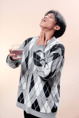 Gri baklava desenli süveter giyen Asyalı bir adam boğazına dokunurken bir bardak su tutuyor ve yukarı bakıyor. Susuzluğunu, sağlık konseptini, susuzluğunu ve insan yaşam tarzını ifade ediyor..