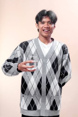 Asyalı adam elinde bir bardak temiz su tutarken gülümsüyor, gri bir baklava desenli süveter giyiyor, su, sağlık, sağlıklı yaşam tarzı, tazelik ve pozitif insan ifadesini temsil ediyor..