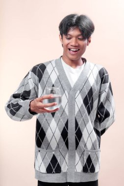 Asyalı bir adam, elinde gri bir baklava desenli süveter, su, sağlık, pozitif yaşam tarzı, tazelik ve enerjiyi sembolize eden bir bardak taze suya mutlu bir şekilde bakıyor..