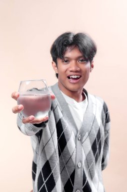 Asyalı bir adam, elinde temiz bir bardak içme suyu tutarken, kendine güvenerek gülümsüyor, şık gri bir baklava desenli süveter giyiyor, neşeli ve pozitif görünüyor..