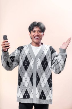 Bir elinde akıllı bir telefonla gülümseyen Asyalı bir adam diğer elinde gri bir baklava desenli süveter, neşe ve güven gösteren bir jest yapıyor..
