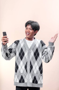 Bir elinde akıllı telefon tutarken gülümseyen Asyalı bir adam diğer elinde gri bir baklava desenli süveter giyiyor, neşe, güven ve modern yaşam tarzını gösteriyor..