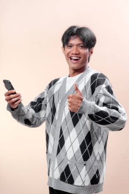 Asyalı bir adam akıllı telefonu tutarken güvenle gülümsüyor ve baş parmak işareti yapıyor gri bir baklava desenli süveter giyiyor, başarıyı temsil ediyor, pozitiflik, iletişim ve yaşam tarzı..