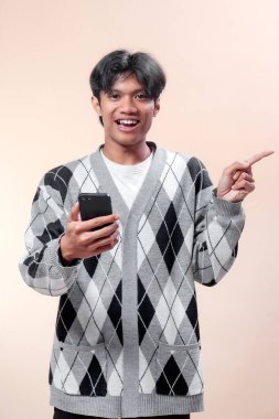 Akıllı telefonu tutarken gülümseyen ve parmağını yana doğru gösteren, baklava desenli süveter giyen neşeli bir yüz ifadesi ve kendine güveni olan Asyalı bir adam..