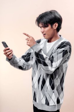 Asyalı bir adam şaşırmış bir ifadeyle akıllı telefona bakıyor, parmağını ekrana doğrultuyor, desenli bir kazak giyiyor, merakını gösteriyor..