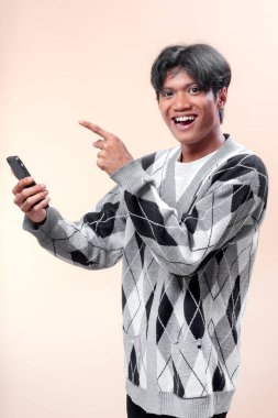 Akıllı telefonu tutarken gülümseyen Asyalı bir adam parmağını yukarı kaldırıyor, baklava desenli süveter giyiyor, heyecan ve kendine güven gösteriyor..