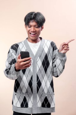 Akıllı telefonu tutarken gülümseyen ve parmaklarıyla yanlamasına gösteren, baklava desenli süveter giyen neşeli ruh hali ve kendine güven gösteren Asyalı bir adam..