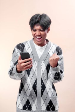 Asyalı adam akıllı telefonu tutarken heyecanlı görünüyor, neşeli bir ifadeyle yumruğunu kaldırıyor, gri baklava desenli süveter giyiyor, mutluluk, başarı ve pozitif enerji gösteriyor..