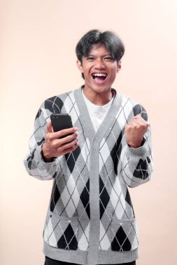 Asyalı adam bir elinde akıllı telefon tutarken parlak bir şekilde gülümsüyor ve yumruğunu heyecanla kaldırıyor, desenli süveter giyiyor, neşe, başarı, kendine güven ve pozitiflik gösteriyor..